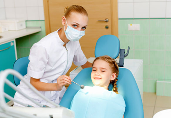 Young girl sitting in a dentist chair, smiling.  The female dentist is holding an inspection mirror towards the girls mouth.